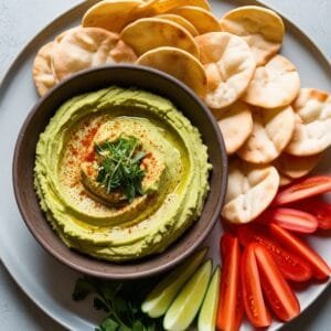 A beautifully plated vegan dish of creamy avocado hummus in a rustic bowl, topped with paprika and fresh herbs, accompanied by crispy pita chips and sliced vegetables for dipping.