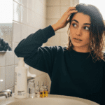 Ultra-realistic editorial photo, mid-shot of a young woman with Mediterranean features in a bright but modest Maltese bathroom, caught mid-motion running her fingers through damp, shoulder-length hair that looks half-frizzy on one side and noticeably smoother on the other, droplets of water clinging to the strands. On the sink in the foreground, slightly out of focus, stands a sleek, unbranded white shampoo bottle with a subtle scientific, clinical design, next to a small cluster of lab-style glass vials containing clear and pale golden liquids, hinting at peptides and active ingredients. The woman’s expression is thoughtful and a little skeptical, as if weighing whether this “science-backed” product really works, while warm Mediterranean daylight spills in from a small window, creating soft highlights on her hair and gentle lens flare at the frame’s edge. The background shows blurred bathroom tiles and a fogged mirror, with faint reflections adding depth. Framing is candid and documentary-style, not posed, as if a moment was captured just as she paused during her routine. Slight motion blur on her moving hand, visible grain equivalent to ISO 800–1600, and subtle imperfections like uneven lighting and minor reflections on the glass create a natural, filmic look. Shallow depth of field keeps focus on her face and hair texture, with the background softly out of focus. No large text elements. COSRX Peptide-132